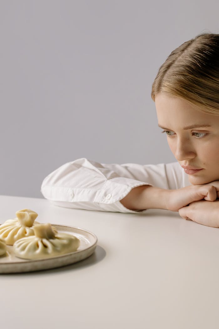 A woman in a white shirt thoughtfully gazes at a plate of dumplings indoors.